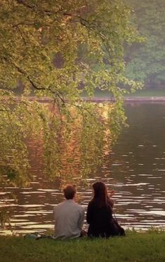 two people are sitting on the grass by the water looking out at the trees in the distance