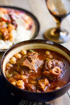 a bowl filled with stew and rice on top of a wooden table next to a glass of wine