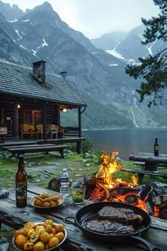 a table with food and drinks on it near a fire pit in front of a cabin