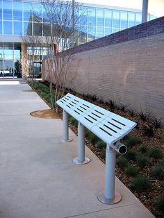 a white bench sitting next to a brick wall near grass and trees in front of a building