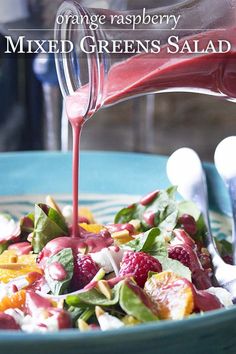 orange raspberry mixed greens salad is being poured into a blue bowl with spoons