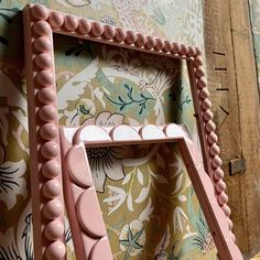 a pink chair with beaded trim around the legs and back rests against a floral wallpapered background