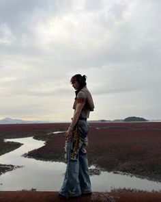 a woman standing on top of a dirt field next to a body of water with mountains in the background