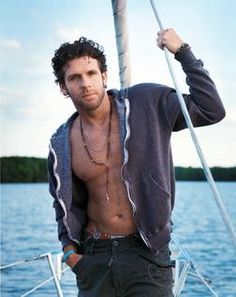 a shirtless man standing on the bow of a sailboat with his hands in his pockets