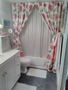 a white bathroom with pink flowers on the shower curtain and rugs in front of the tub