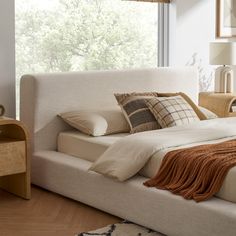 a bed with pillows and blankets on top of it next to a night stand in front of a window