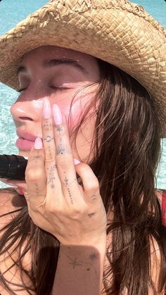 a woman wearing a straw hat and holding her hand up to her face while standing in front of the ocean