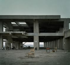 two wooden chairs sitting in front of a large building with concrete columns on the roof