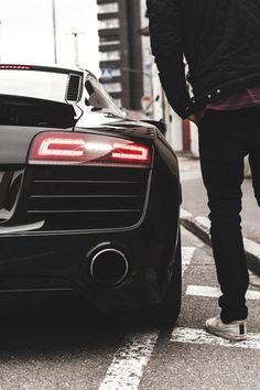 a man walking across a street next to a black sports car with its tail lights on