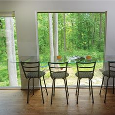 a glass table with four chairs in front of a large window that looks out onto the woods