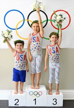 three young children standing on pedestals holding flowers in front of the olympic rings wall