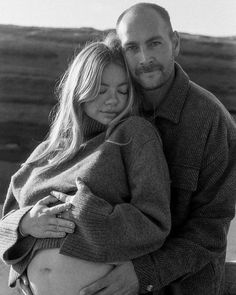 black and white photograph of a pregnant couple hugging each other in front of the ocean