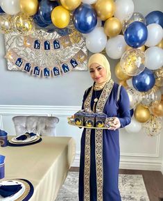 a woman holding a cake in front of a table with blue and gold balloons on it