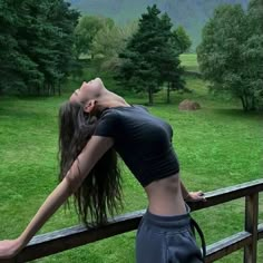 a woman leaning on a wooden fence with her arms behind her head and trees in the background