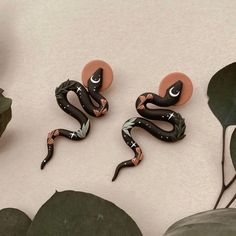 two black and orange snake earrings sitting on top of a white table next to plants