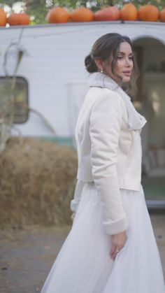 a woman in a white dress and jacket walking down the street with pumpkins behind her