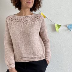 a woman standing in front of a white wall wearing a pink sweater and black pants