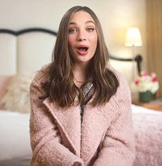 a woman in a pink coat is sitting on a bed and making a surprised face