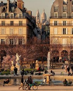 many people are sitting and walking around in the park near some buildings with water fountains