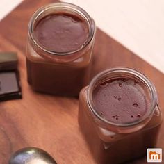 two jars filled with chocolate sitting on top of a wooden table next to a spoon