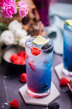 two blue drinks with raspberries and lime on the rims, one has a lemon wedge in it