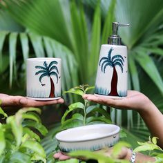 two hands holding cups with palm trees painted on them in front of green plants and foliage