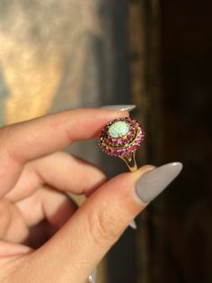 A delicious mid-century adornment, with a luminescent dazzle! Crafted circa 1970’s in 18 carat gold, this Opal and Ruby ring is a vibrant cluster of sparkles! No matter how you wear this ring, it is a feast for the eyes. The crackling colors of this exceptional opal pick up beautifully against the vibrant jam tones of the double ruby halo. Double prongs complete the regal look of the ring, creating a sophisticated yet timeless silhouette. One of the loveliest details is the undercarriage; with its scrolling foliate design and slight open back, it is a wonderful example of the classic 1970’s ring design. A wonderful vintage treasure, perfect for the antique connoisseur and romantic alike! Materials and Features: 18ct, Opal, Ruby Marks: 18K Age/Origin: 1970’s, American Measurements: Size 5, Yellow Gold Multi-stone Rings For Party, Dazzling Oval Multi-stone Cluster Ring, Dazzling 17 Jewels Cluster Ring, Vintage Yellow Gold Halo Jewelry, Yellow Gold Cluster Ring With Gemstone, Vintage Halo Cluster Ring Gift, Vintage Cluster Ring With Halo As Gift, Vintage Halo Cluster Ring As Gift, Beautiful Rings Vintage