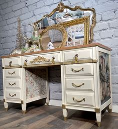 an antique white desk with gold trimming and ornate mirror on top, against a brick wall