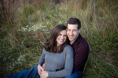 a man and woman sitting on the ground in front of tall grass with wildflowers