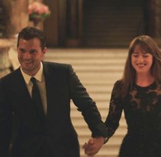 a man and woman holding hands walking down a hall way in formal wear, smiling at the camera