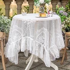 a white table topped with a vase filled with flowers next to two chairs and a small potted plant