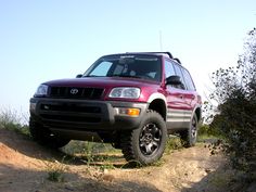 a red four door suv parked on top of a dirt hill next to bushes and trees