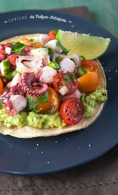 an open pita topped with vegetables and meat on top of a blue plate next to a lemon wedge