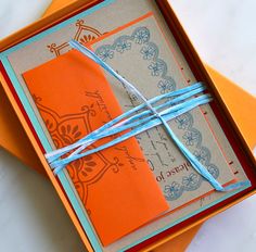 an orange and blue wedding card in a box with ribbon tied around the edge on top of it