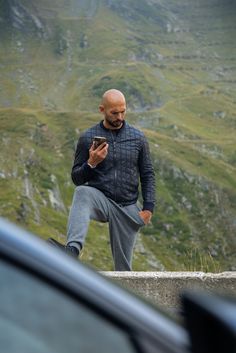 a bald man standing on top of a hill looking at his cell phone while wearing sweatpants