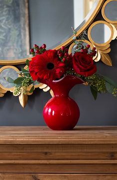 a red vase filled with flowers sitting on top of a wooden table next to a mirror