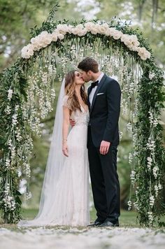 a bride and groom kissing in front of an arch decorated with white flowers, greenery and foliage