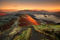 the sun is setting on top of a mountain with a lake in the foreground