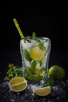 a glass filled with ice and limes on top of a black table next to two lemon wedges