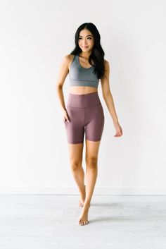 a woman standing in front of a white wall wearing purple shorts and a gray tank top