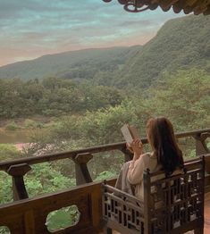 a woman is sitting on a bench looking out at the mountains and trees while reading a book