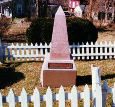 a monument in the middle of a yard with a white picket fence around it and a house behind it