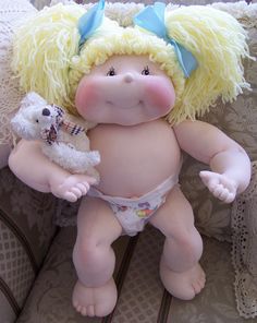 a baby doll with blonde hair holding a teddy bear and wearing a diaper on a couch