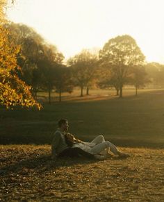 two people sitting on the ground in a field with trees and sun shining behind them