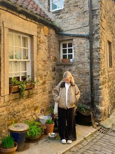 a woman standing in front of a stone building with potted plants on the sidewalk