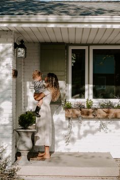 a woman holding a child in front of a white house with potted plants on the porch