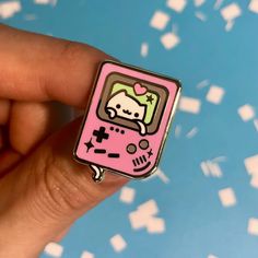 a person is holding a pink gameboy pin in their left hand, with white letters on the background