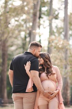 a pregnant woman standing next to a man with her belly wrapped around him in front of trees