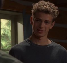 a young man with curly hair standing in front of a window looking at another person