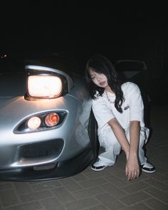 a woman leaning against the hood of a sports car at night with her hands on the door handle